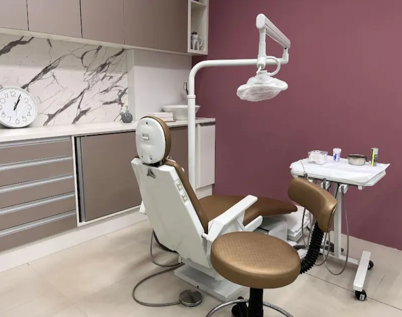 Sala de atendimento com cadeira odontológica bege, refletor, armários claros e parede em tom rosado