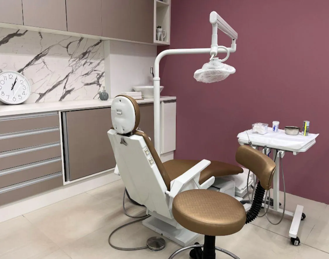 Sala de atendimento com cadeira odontológica bege, refletor, armários claros e parede em tom rosado